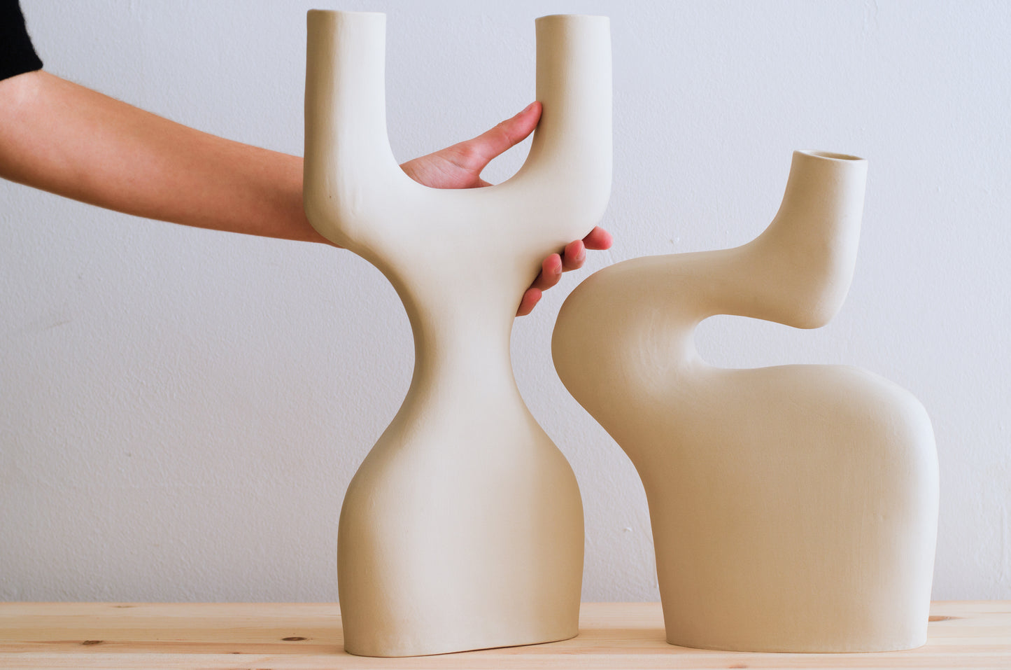 Una mano di una ragazza si appoggia sopra un vaso di ceramica fatto a mano. Il vaso ricorda la forma di una Y ma a base larga ed è a fianco di un altro vaso dalle forme sinuose. I due vasi hanno le curve che si incastrano e si completano. I vasi sono tutti e due beige e sono fatti a mano.