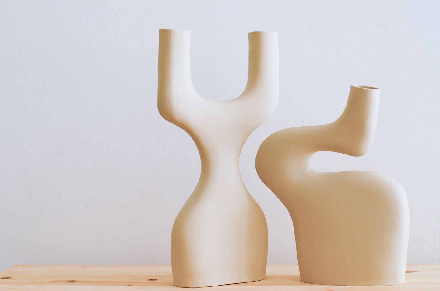 Nella foto ci sono due vasi di ceramica fatti a mano. Un vaso ricorda la forma di una Y ma a base larga ed è a fianco di un altro vaso dalle forme sinuose. I due vasi hanno le curve che si incastrano e si completano. I vasi sono tutti e due beige e sono fatti a mano.