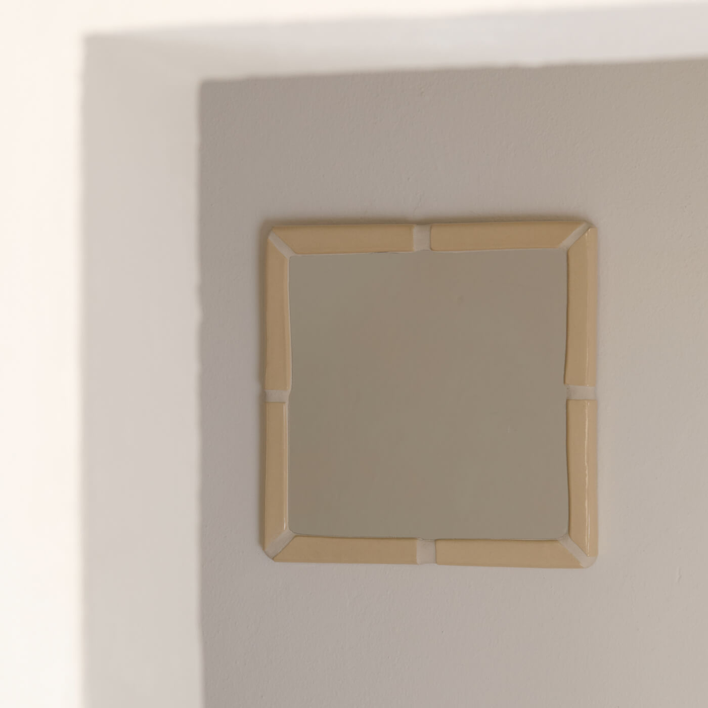 Specchio quadrato Kalakera con cornice in ceramica artigianale panna, illuminato da una luce radente che ne esalta i volumi. Un complemento di design materico per un arredamento minimalista e ricercato.
