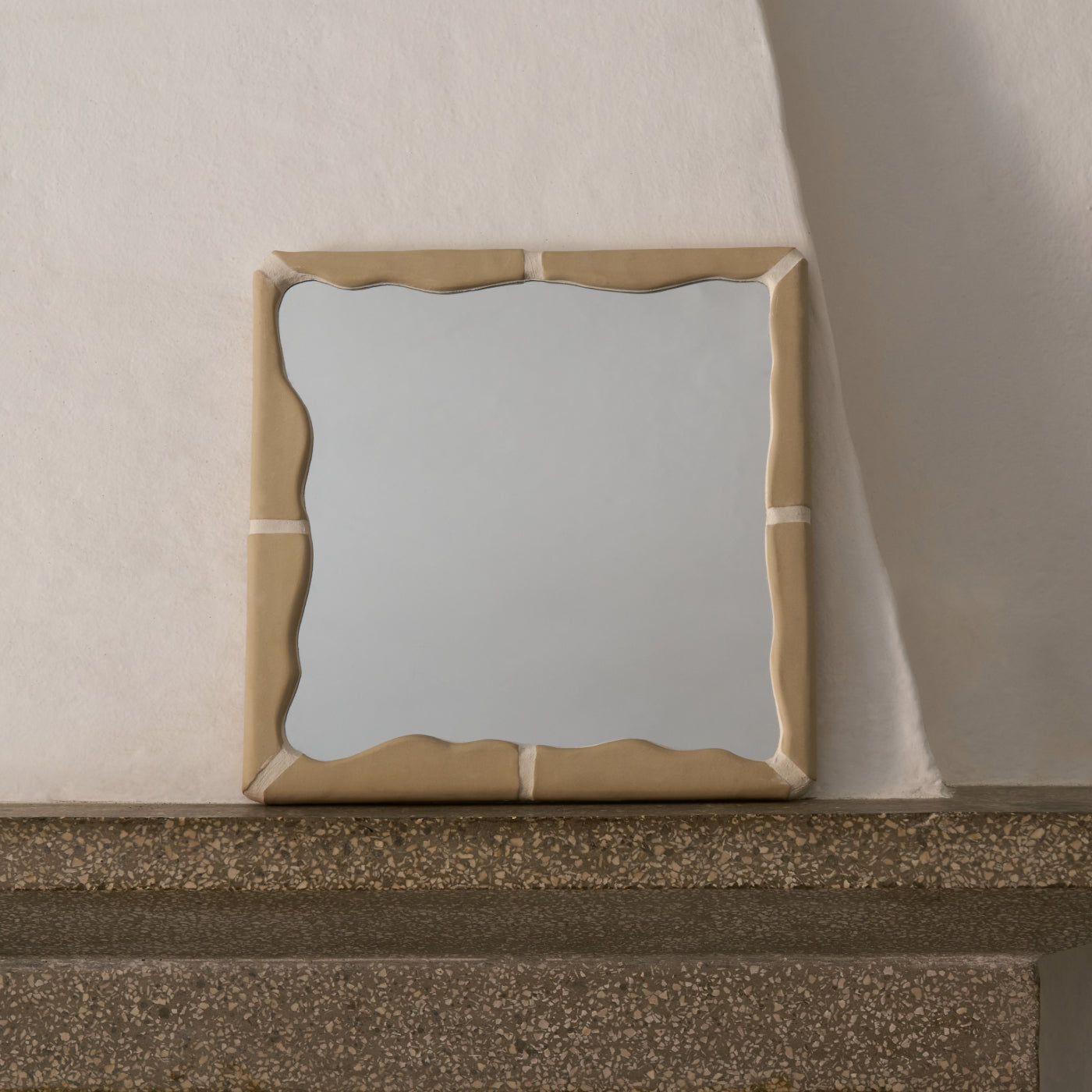 Specchio quadrato Kalakera con cornice in ceramica artigianale color terra, appoggiato su un ripiano in pietra. Il design a onde e la texture materica lo rendono un elemento scultoreo ideale per interni in stile mediterraneo moderno.