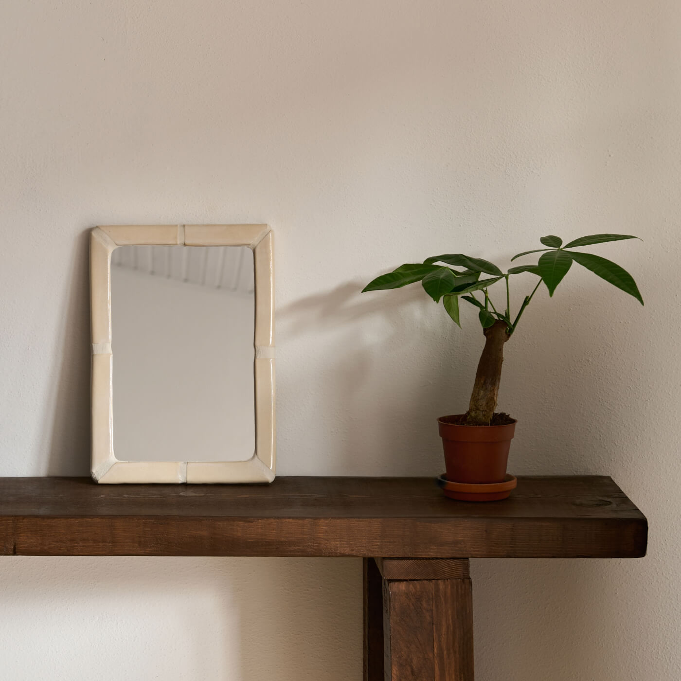 Specchio rettangolare Kalakera con cornice in ceramica artigianale color panna, appoggiato su una console in legno massiccio accanto a una pianta verde. Un esempio di design materico e minimale per ingressi raffinati.