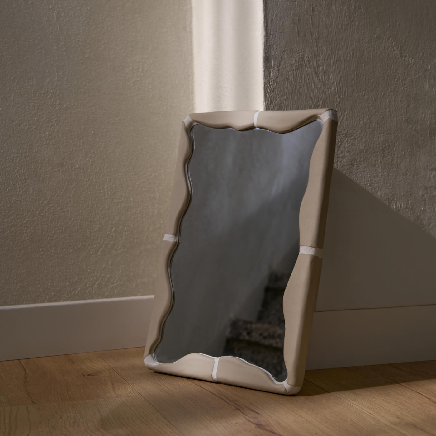Specchio rettangolare Kalakera con cornice in ceramica fatta a mano color beige, appoggiato a terra in un interno dal design minimale. La cornice presenta una silhouette scultorea a onde che enfatizza l'unicità del pezzo artigianale.