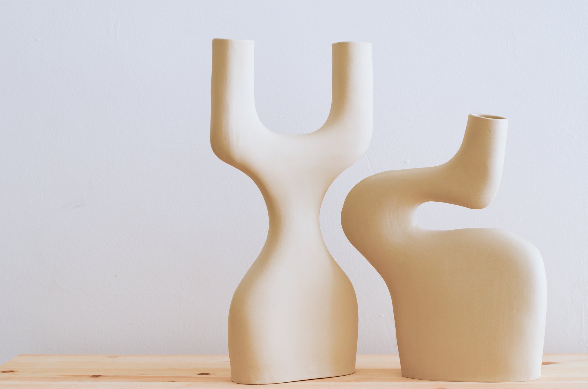 Nella foto ci sono due vasi di ceramica fatti a mano. Un vaso ricorda la forma di una Y ma a base larga ed è a fianco di un altro vaso dalle forme sinuose. I due vasi hanno le curve che si incastrano e si completano. I vasi sono tutti e due beige e sono fatti a mano.