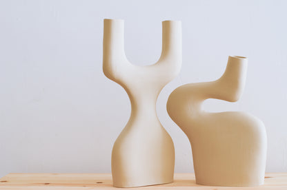 Nella foto ci sono due vasi di ceramica fatti a mano. Un vaso ricorda la forma di una Y ma a base larga ed è a fianco di un altro vaso dalle forme sinuose. I due vasi hanno le curve che si incastrano e si completano. I vasi sono tutti e due beige e sono fatti a mano.